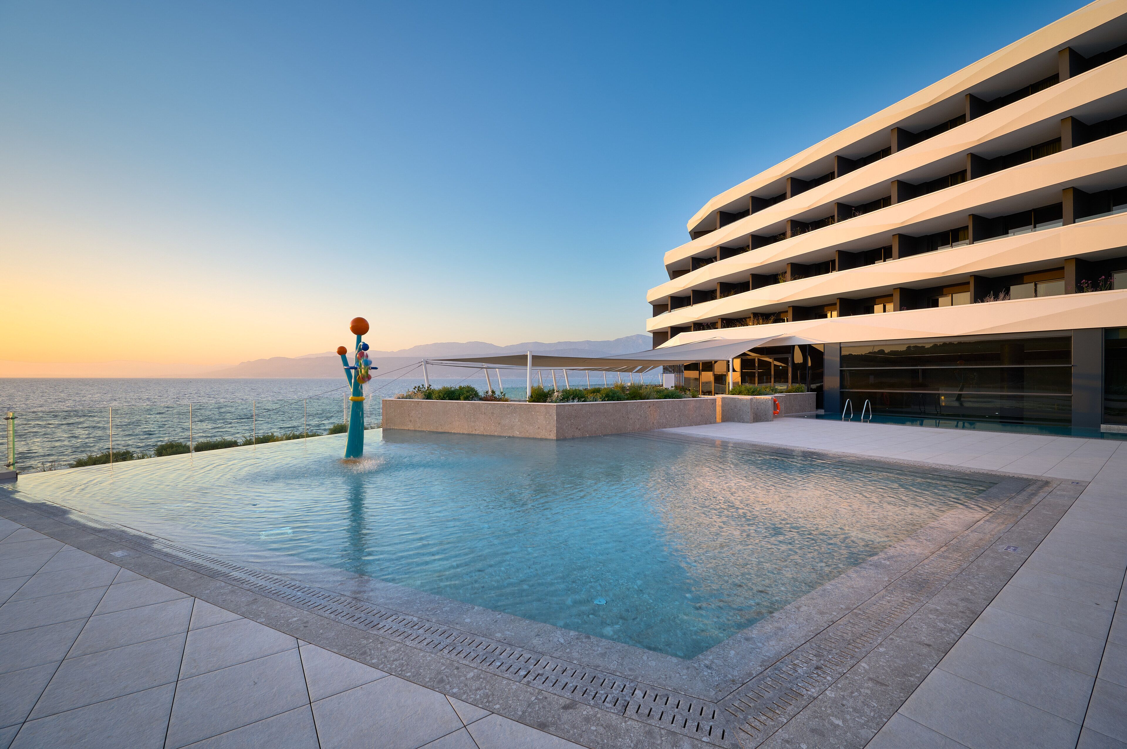 Un hôtel moderne doté d’une piscine avec vue sur la mer, caractérisé par une structure à plusieurs étages et un espace salon couvert.