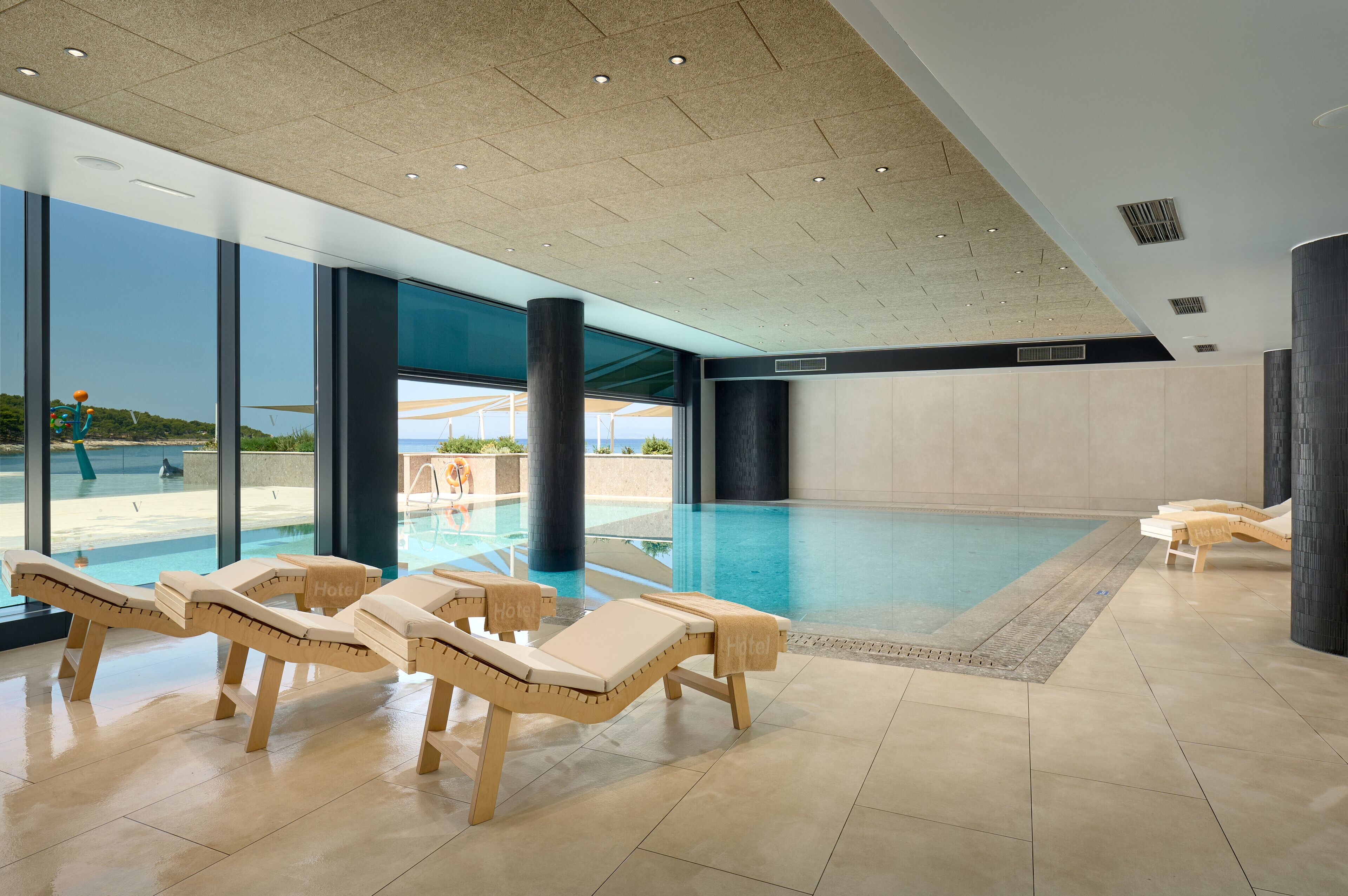 A modern indoor swimming pool with lounge chairs and large windows offering views of the beach.