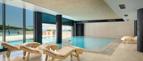 A modern indoor swimming pool with lounge chairs and large windows offering views of the beach.