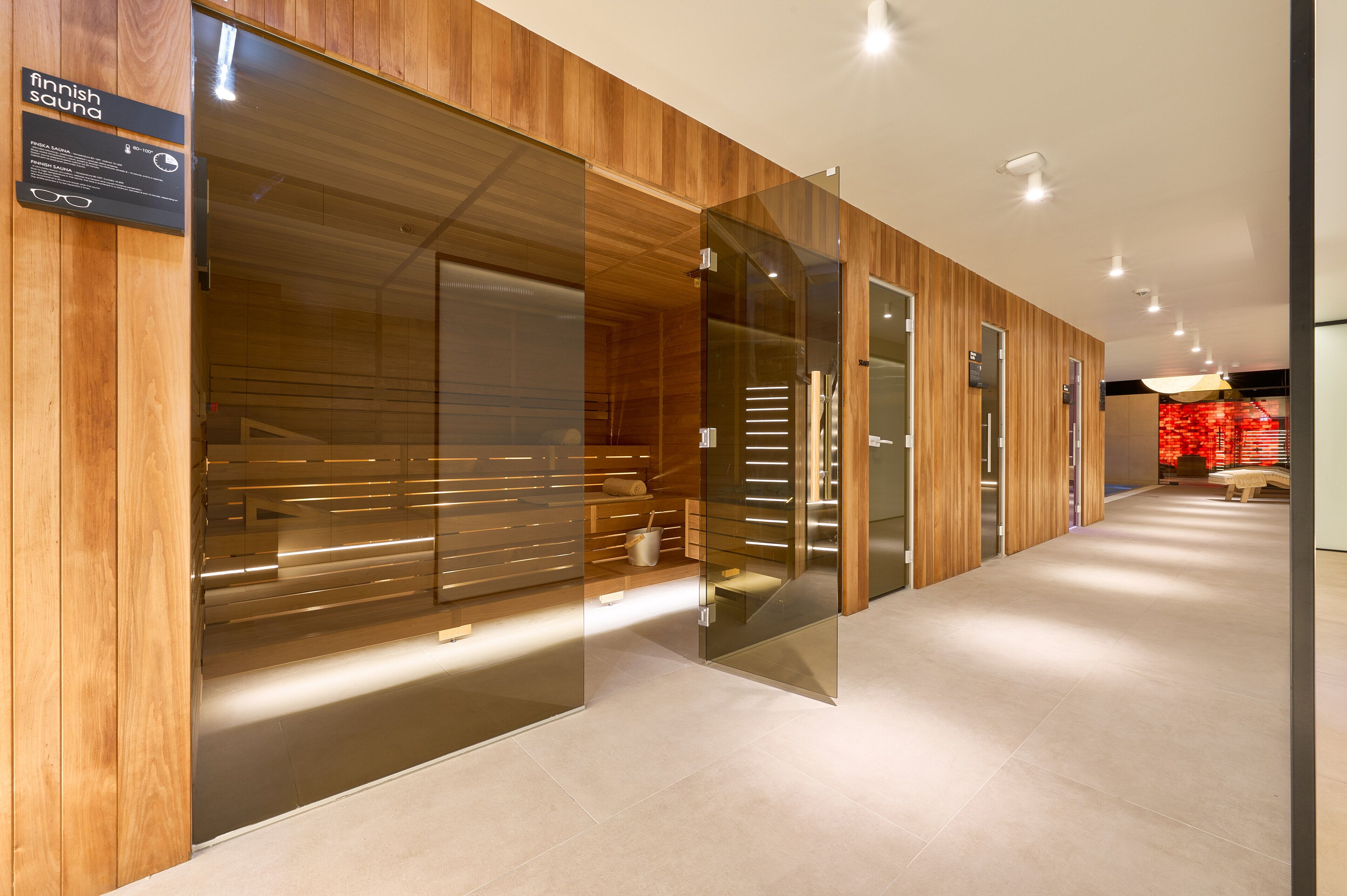 Eine moderne Sauna mit Holzverkleidung und Glastüren, ein Schild weist auf eine finnische Sauna hin, und ein gefliester Boden.