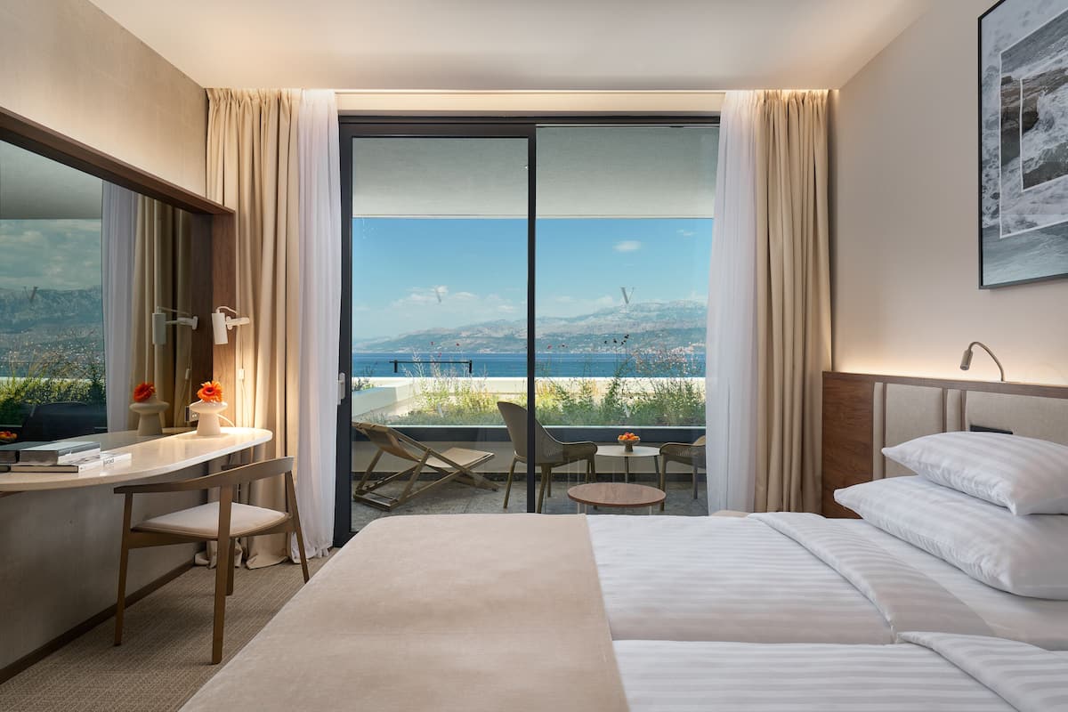 A hotel room with a bed, a desk, a chair, a small table, and a view of the sea.