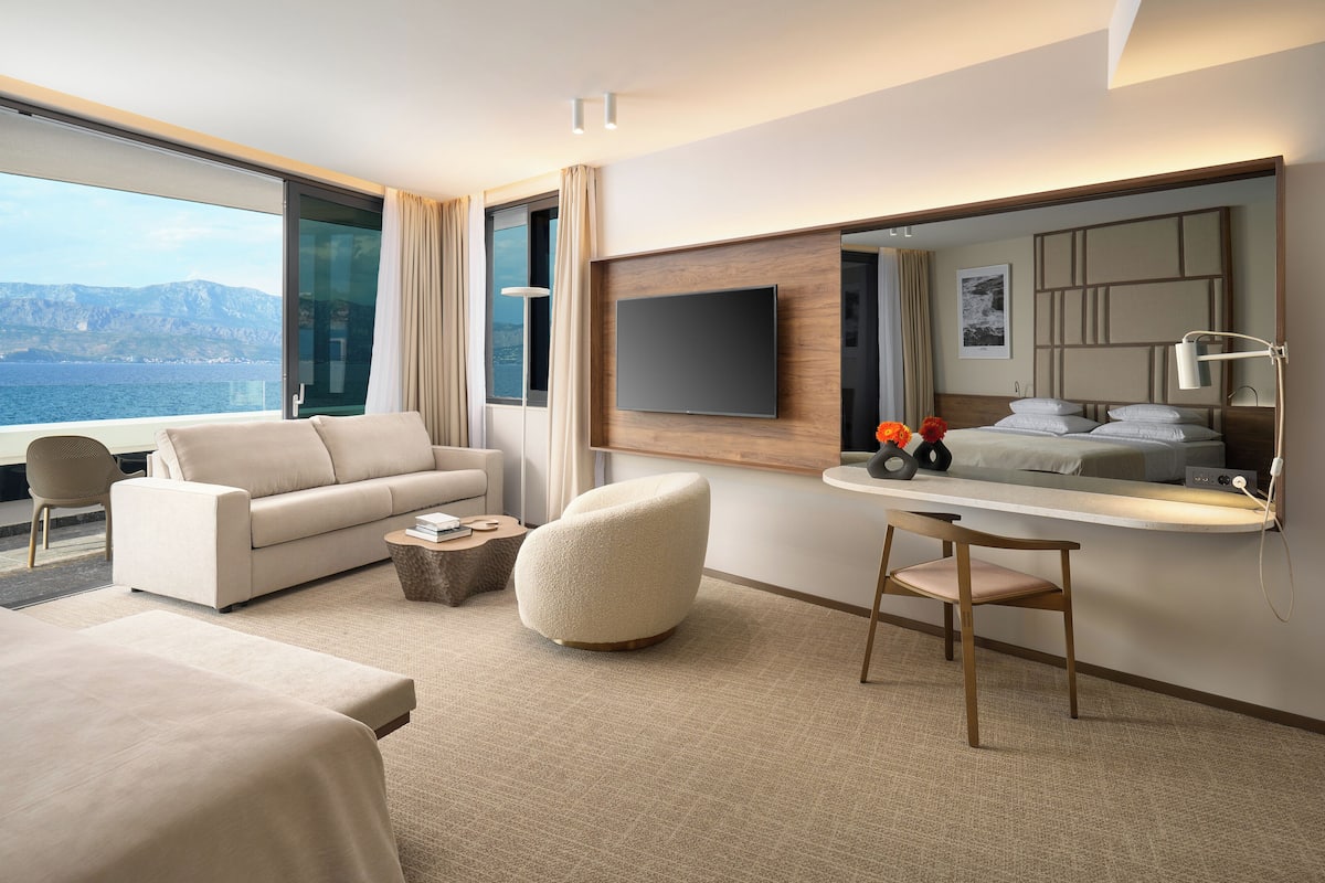 A modern hotel room with a large window offering a sea view, a sofa, a round ottoman, a small table with books, and a flat-screen TV mounted on the wall.