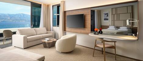A modern hotel room with a large window offering a sea view, a sofa, a round ottoman, a small table with books, and a flat-screen TV mounted on the wall.