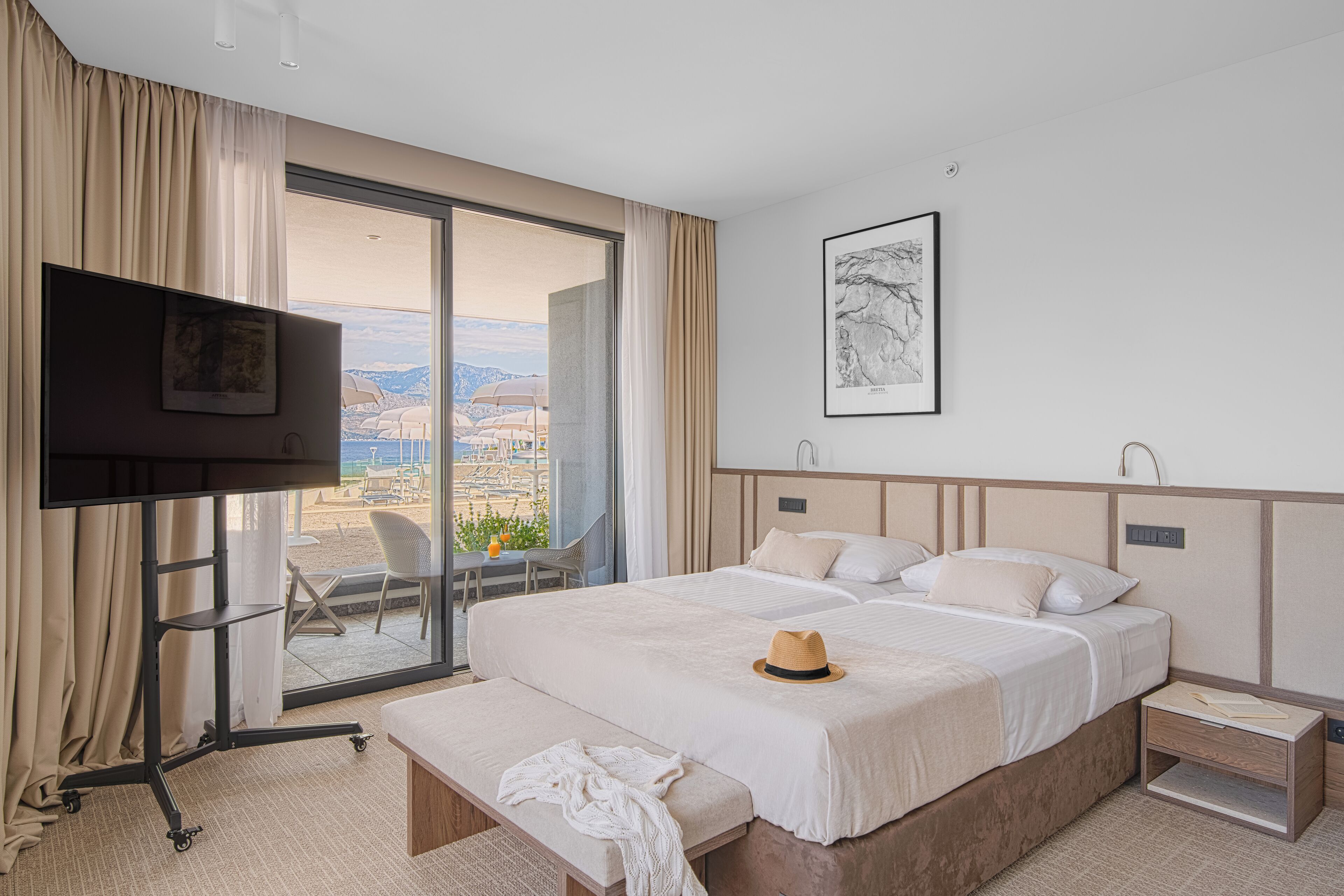 A hotel room with a large bed, a TV, a desk with a chair, and a balcony with a view of the city.