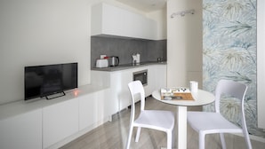 A modern, minimalist kitchenette with a flat-screen TV, a coffee maker, and a kettle. There is a small dining area with two white chairs and a table, and a magazine on the table. - Italianway - The B Village Business - 12 (Milan)