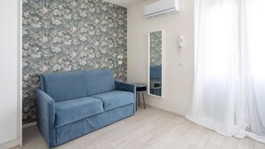 A room with a blue sofa, a small round table, a mirror, and a white curtain. - Italianway - The B Village Superior - 40 (Milan)