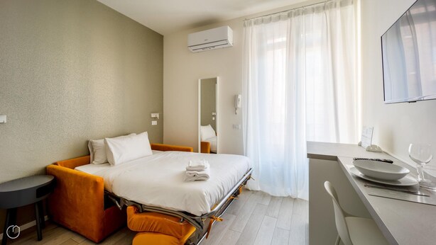 A hotel room with a bed, a desk, a chair, an air conditioning unit, and a window with curtains. - Italianway - The B Village Business - 30 (Milan)