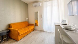 A modern, well-lit room with a sofa, a small table, a mirror, and a kitchen area with a counter and a wine glass. - Italianway - The B Village Business - 30 (Milan)