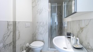 A modern bathroom with a marble floor, a white toilet, a glass-enclosed shower, and a large sink with toiletries. - Italianway - The B Village Business - 34 (Milan)