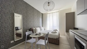 A modern hotel room with a large bed, a desk with a chair, a small table, a mirror, and a kitchenette. - Italianway - The B Village Business - 34 (Milan)