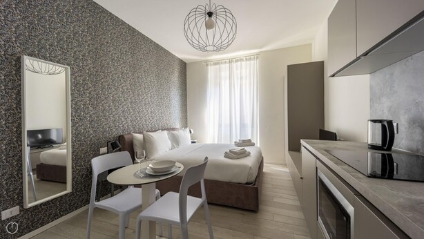 A modern hotel room with a bed, a dining table with chairs, a kitchen area, and a large mirror. - Italianway - The B Village Business - 34 (Milan)