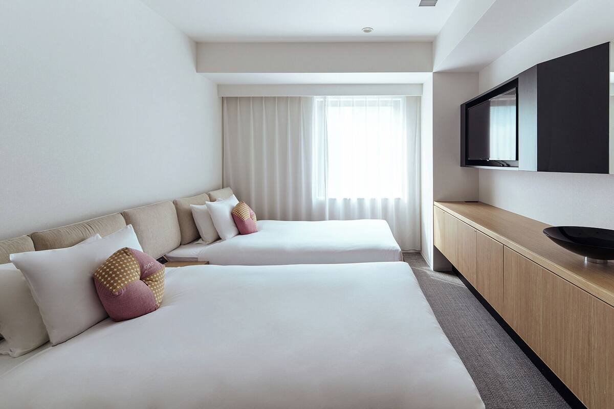 A hotel room with two beds, a TV mounted on the wall, and a large window with curtains.