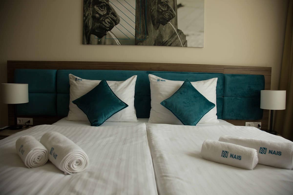 A hotel room with two beds, teal headboard, white linens, teal pillows, and rolled white towels with "NAIS" branding.