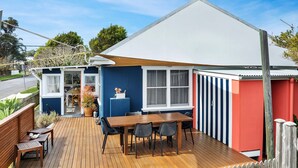 Une petite maison de plain-pied avec une terrasse en bois, un coin repas avec une table et des chaises, et un petit jardin agrémenté d’une plante en pot.