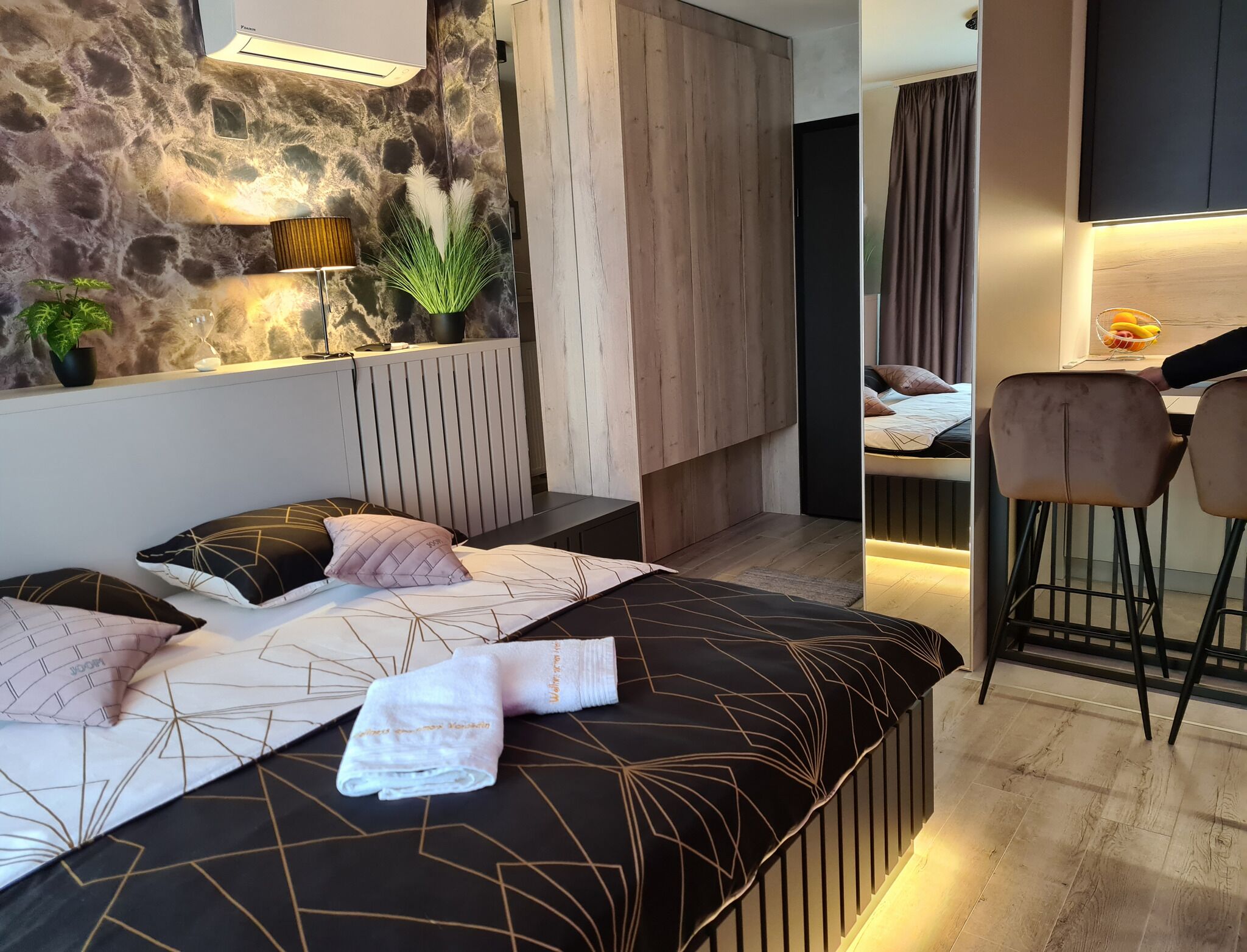 Une chambre moderne avec un mur en pierre, un lit avec une literie à motifs géométriques, une plante sur la table de chevet et un petit coin salon avec une chaise et une table.