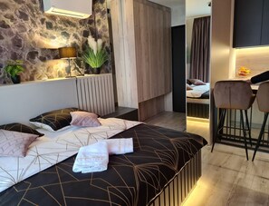 A modern bedroom with a stone wall, a bed with geometric patterned bedding, a bedside plant, and a small seating area with a chair and table. - Wellness Apartments (Varazdin)