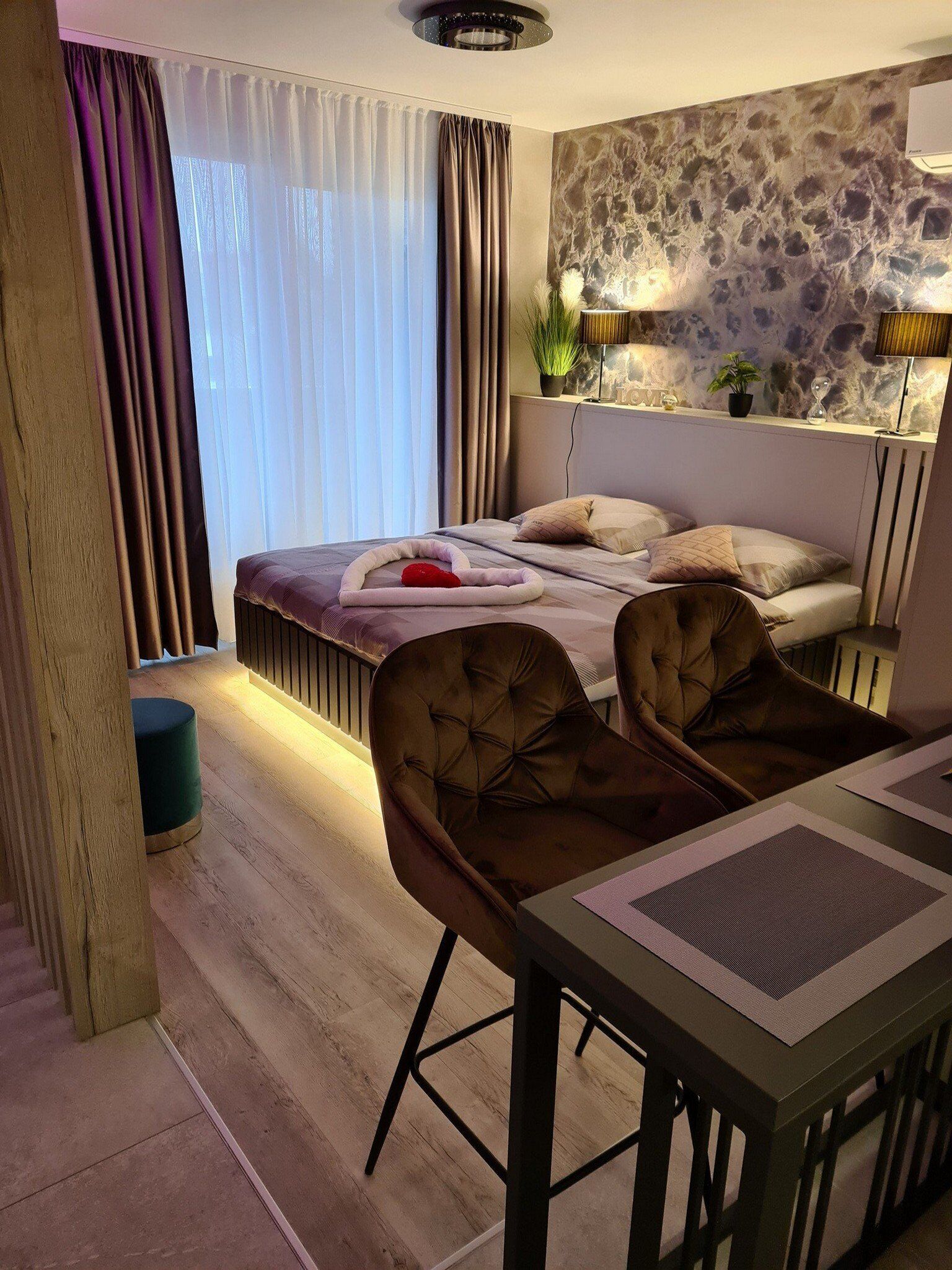 A modern hotel room with a bed, two armchairs, a small table, and a stone wall.