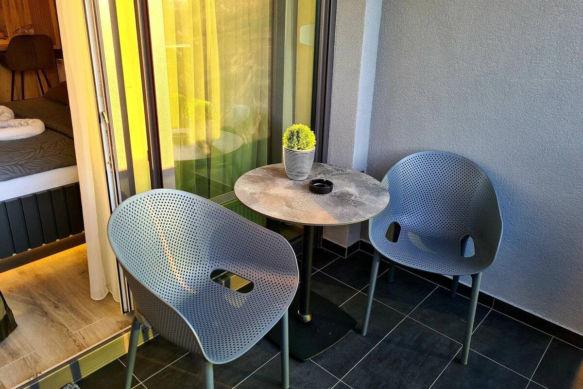 A small outdoor seating area with two perforated metal chairs and a round table, a potted plant, and a glimpse of a room with a bed and a chair.