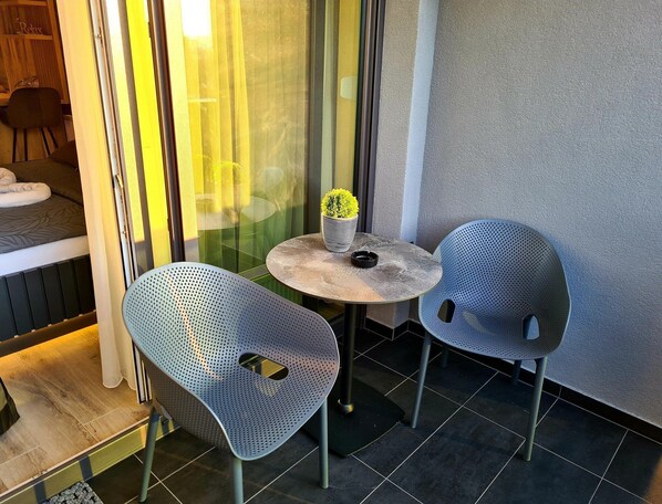 A small outdoor seating area with two perforated metal chairs and a round table, a potted plant, and a glimpse of a room with a bed and a chair. - Wellness Apartments (Varazdin)