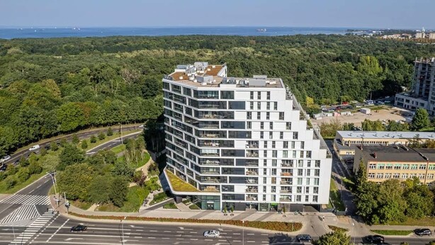 Un bâtiment moderne à plusieurs étages, doté d’un toit plat et de grandes fenêtres, entouré de verdure et bordé par une route.