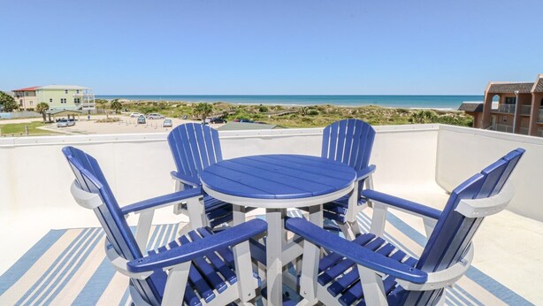 Eine Terrasse mit einem runden Tisch und vier blauen Stühlen, mit Blick auf einen Strand und den Ozean.