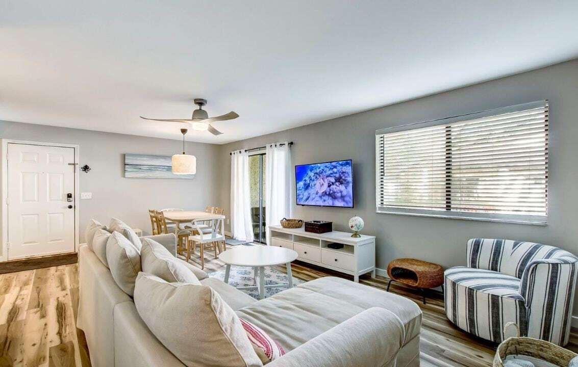Ein Wohnzimmer mit einer Couch, einem Fernseher, einem Deckenventilator, einem Esstisch und einem Fenster mit Jalousien.