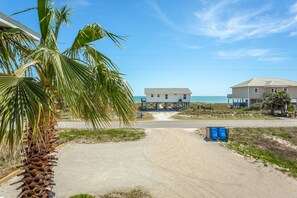 A coastal property with a sandy driveway leading to a house elevated on stilts, a beach view, and a palm tree in the foreground.