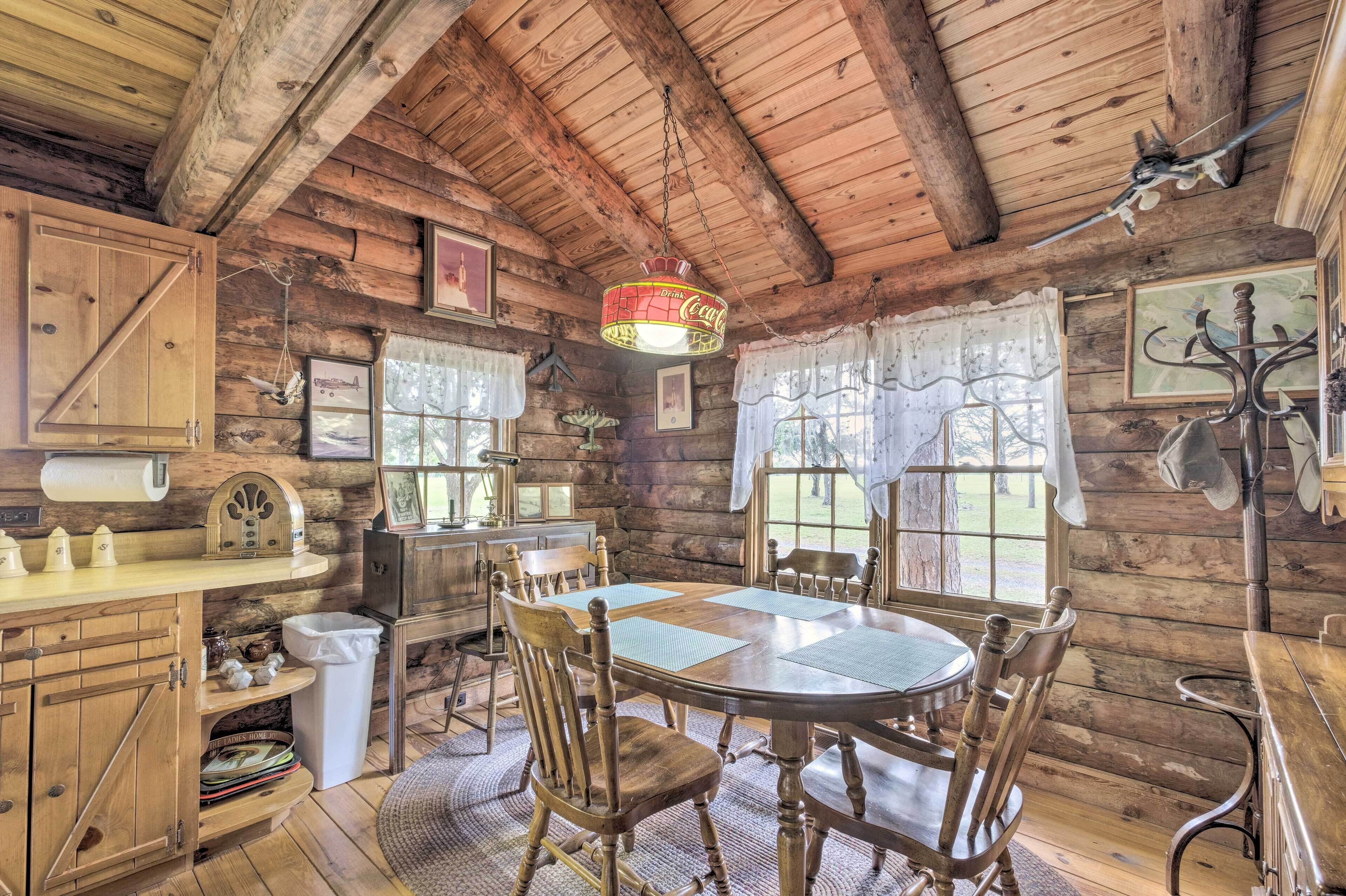 A rustic wooden cabin interior with a dining table, chairs, a hanging Coca-Cola sign, and a large window with curtains.