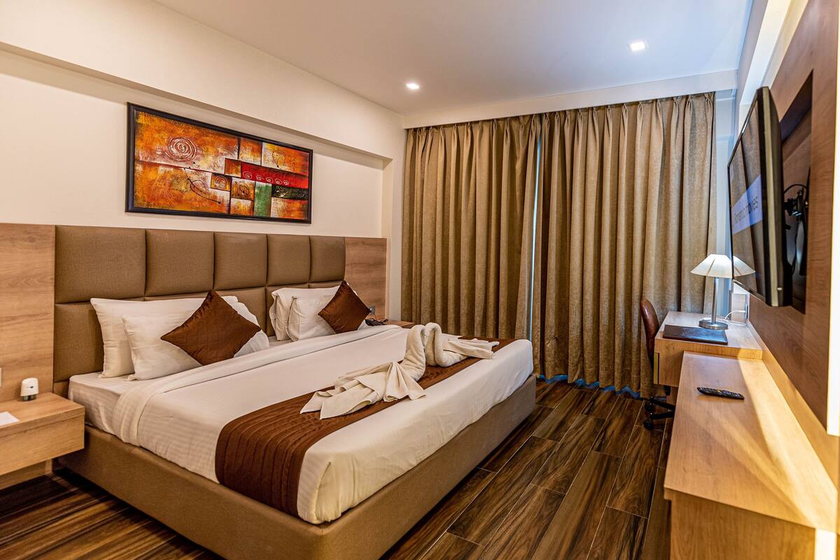 Una habitación de hotel con una cama grande, un cabecero de madera, una mesita de noche con lámpara y un ventanal con cortinas.