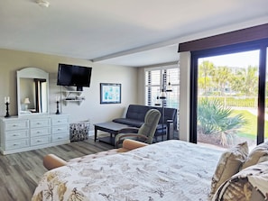 Un dormitorio con cama, sofá, televisor, cómoda y ventana con vista a palmeras.