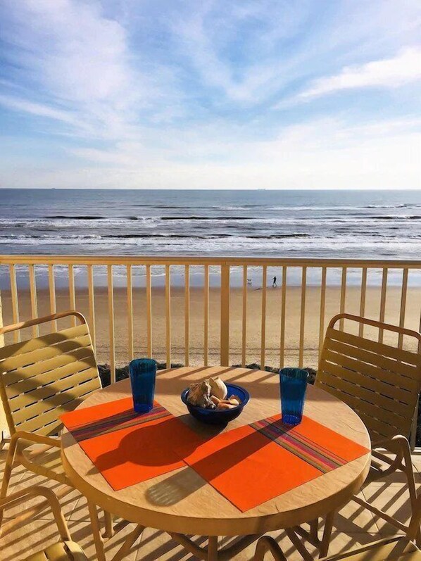 A round table with orange placemats, two blue glasses, and a bowl of snacks on a balcony overlooking the ocean.
