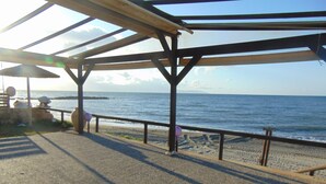 A beachside walkway with a wooden pergola, a thatched umbrella, and a sandy beach with the ocean in the background. - Litinas Casa-In front of the Sea (Chania)