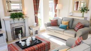 A living room with a fireplace, sofa, and a plaid ottoman. - Curlew Lodge (Carnforth)