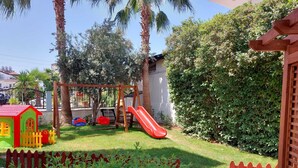 A backyard with a wooden swing set, a red slide, a small playhouse, and a fenced area with greenery. - Summer Rose Hotel (Manavgat)