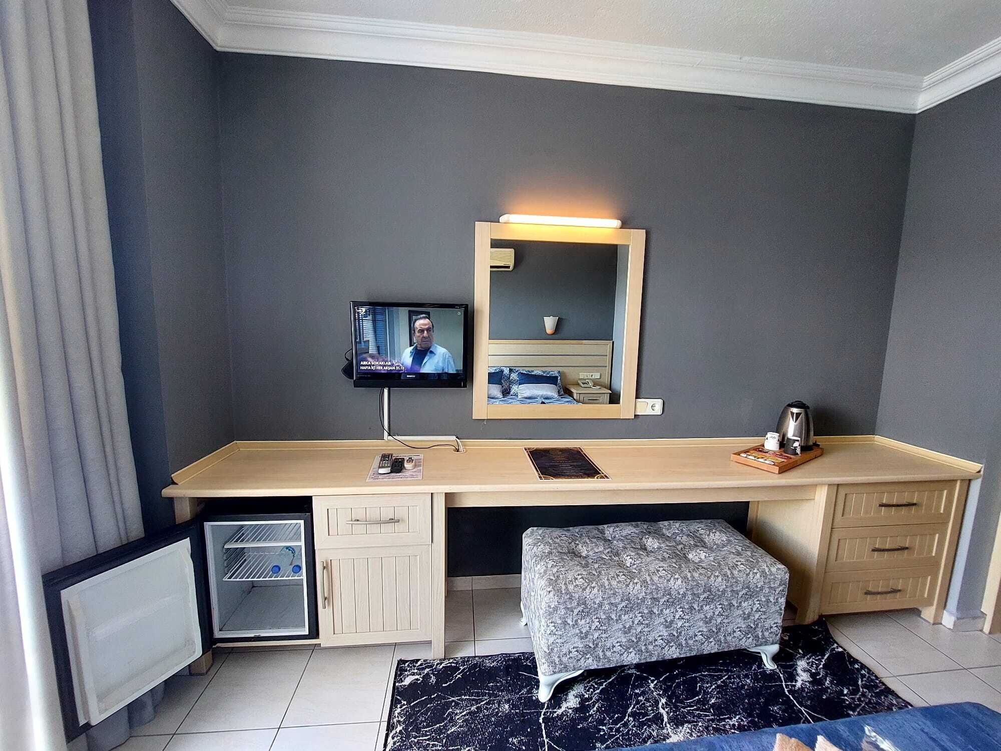 Quarto de hotel com escrivaninha, televisão, cama, pequeno frigorífico, um banco e chão ladrilhado.