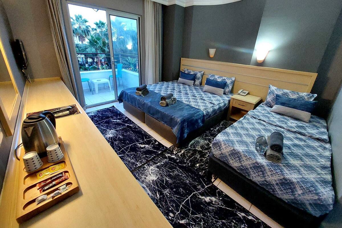 A hotel room with two beds, a small table with a kettle and cups, a nightstand with a phone, and a view of a pool and palm trees through the glass door.