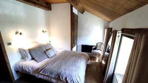 Un dormitorio con techo de madera, una cama con colcha a rayas grises y blancas, dos luces fijadas en la pared, una silla y un televisor.