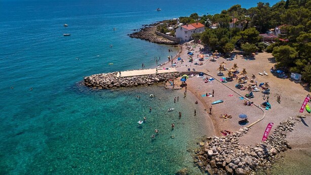 A coastal beach with clear waters, a rocky breakwater, and a row of buildings with red roofs. - Premium Apartment Panoramico – Spacious 4-Star Holiday Apartment with a Sea view (Krk)