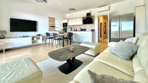 A modern living room with a sofa, a coffee table, and a TV. There is a kitchen area with cabinets and a refrigerator. - Premium Apartment Panoramico – Spacious 4-Star Holiday Apartment with a Sea view (Krk)