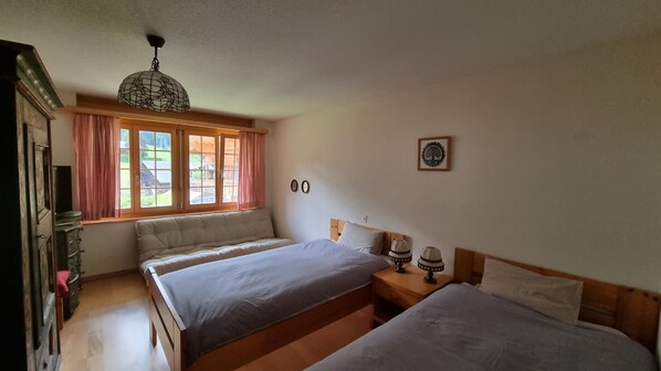 A bedroom with two beds, a wooden dresser, a window with curtains, and a hanging light fixture. - Room with extra-large double-bed, cable TV, private bathroom, organic breakfast, (Lauenen)