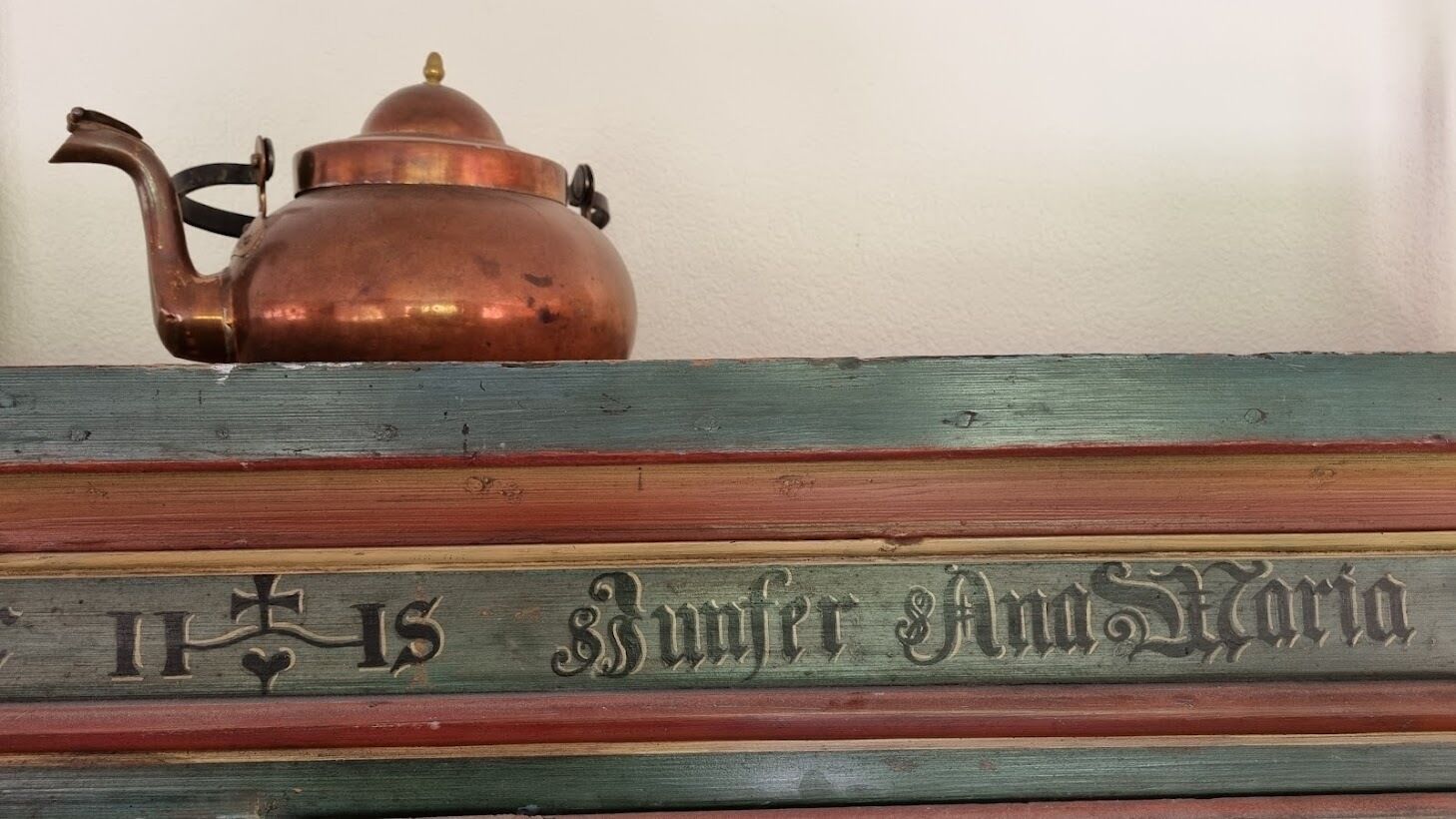 Une théière en cuivre est posée sur une surface en bois, et on aperçoit partiellement la couverture d’un livre intitulé « Buntor et Maria ».