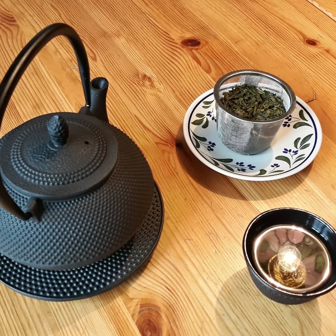 Una tetera negra sobre una mesa redonda de madera, una taza con motivos florales y una taza negra pequeña con una vela encendida.