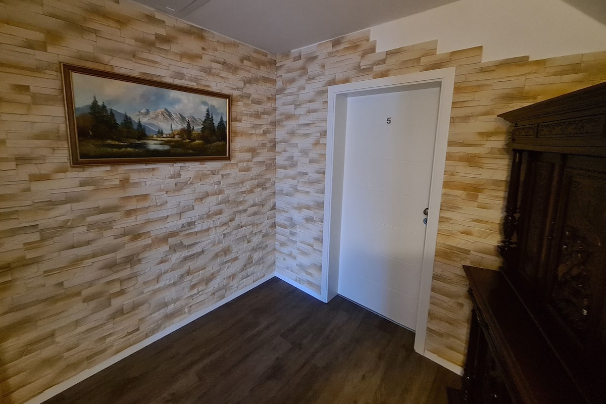Un couloir avec un mur décoré d’un motif imitant la pierre, une porte portant le numéro 5 et un tableau représentant un paysage de montagne.