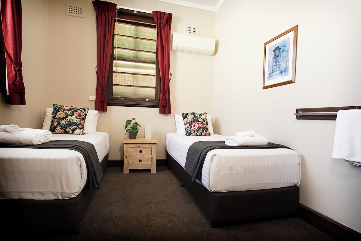 A hotel room with two beds, a small bedside table with a plant, a window with curtains, an air conditioning unit, and a framed picture on the wall.