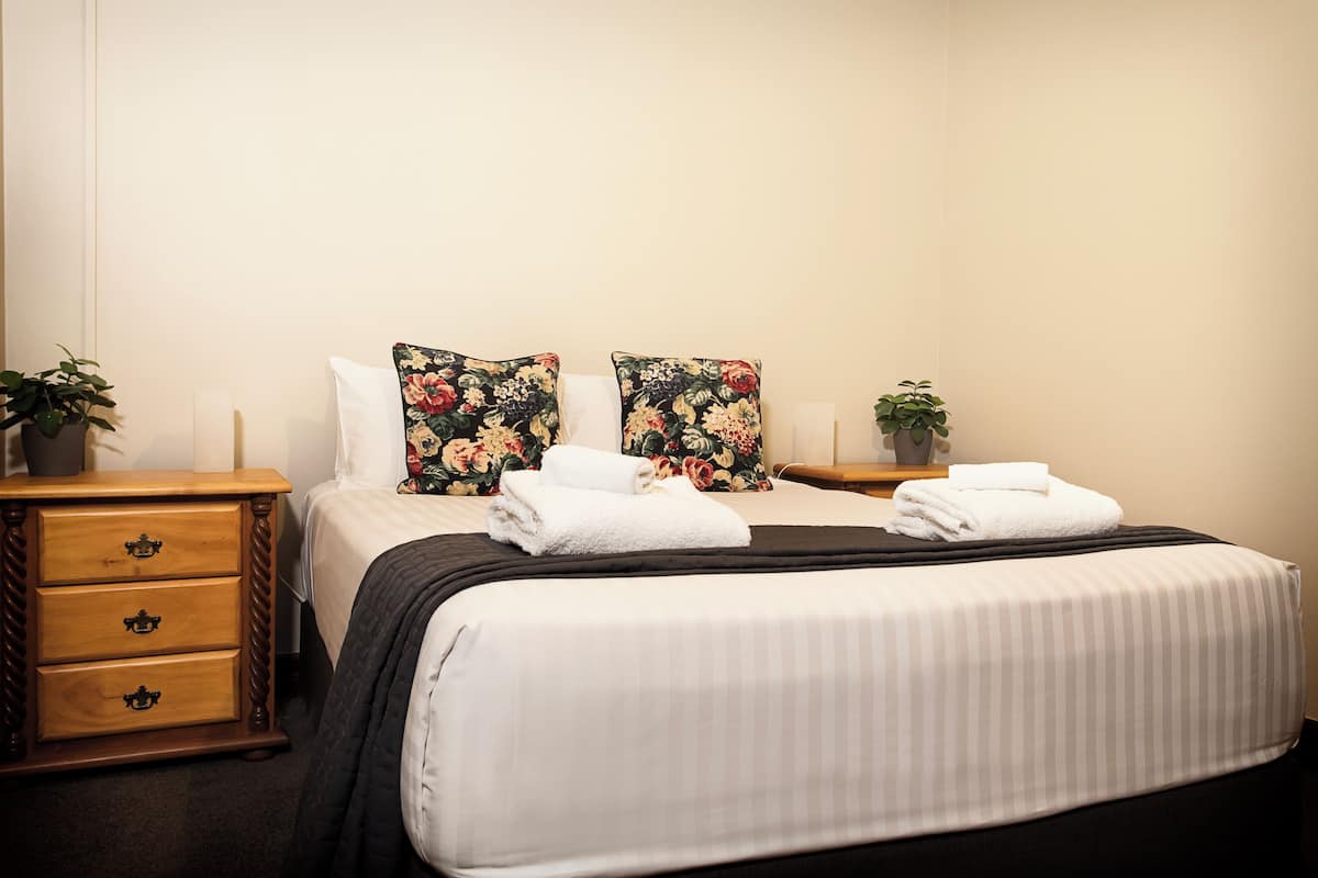 A neatly made bed with white linens and two floral pillows, a wooden nightstand with drawers, and a small potted plant on the nightstand.