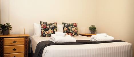 A neatly made bed with white linens and two floral pillows, a wooden nightstand with drawers, and a small potted plant on the nightstand.