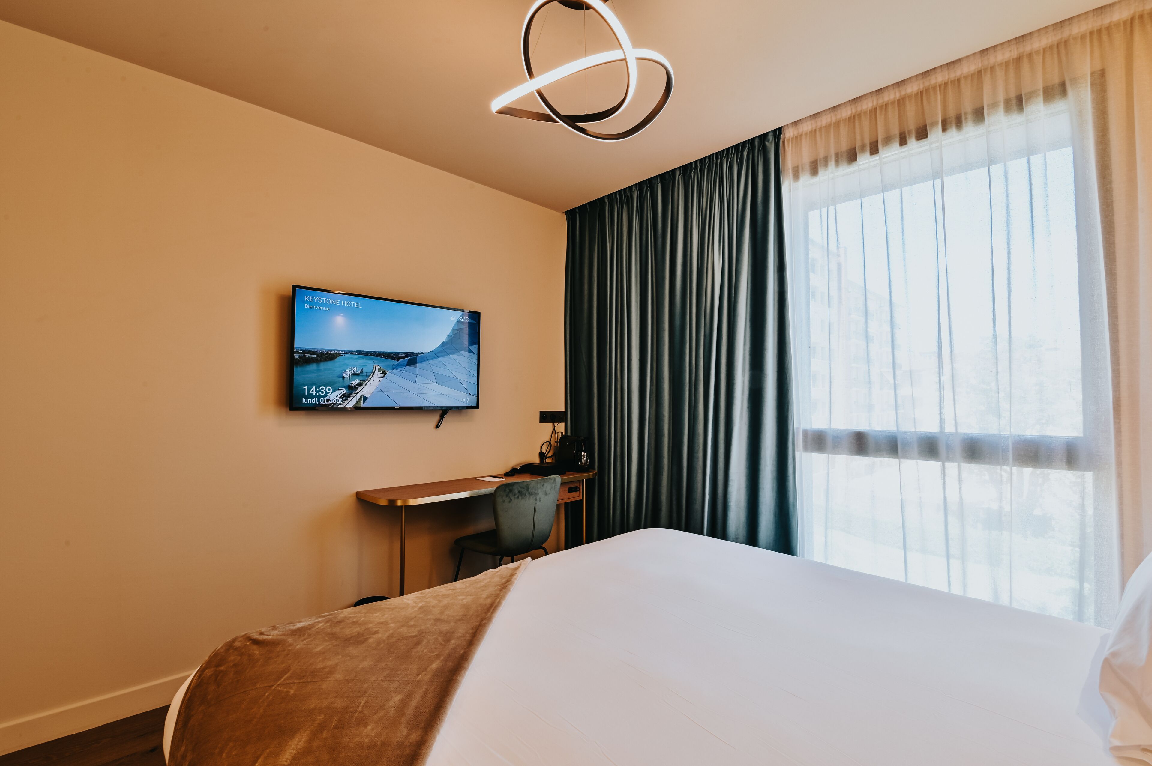 A hotel room with a bed, a desk, a chair, a television mounted on the wall, and a window with curtains.