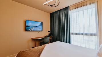 A hotel room with a bed, a desk, a chair, a television mounted on the wall, and a window with curtains.
