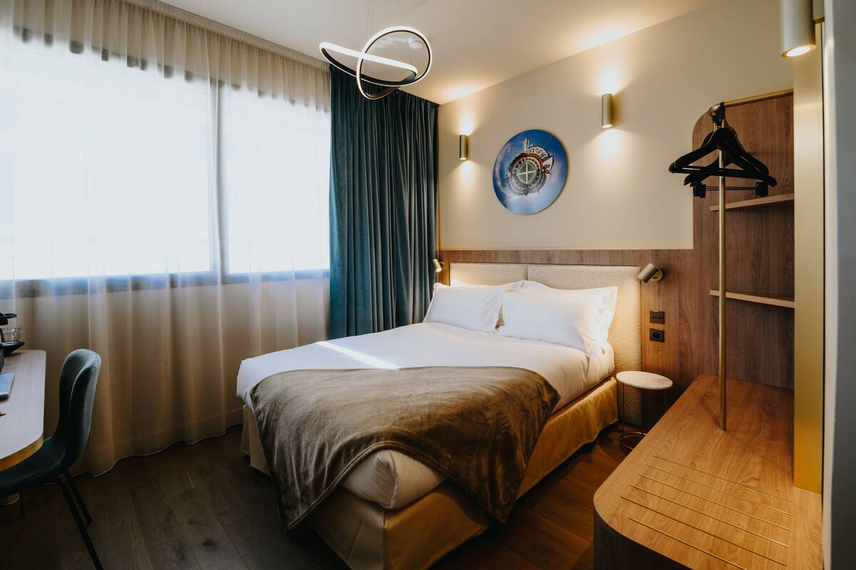 A hotel room with a bed, a desk, a chair, a coat rack, and a wall-mounted clock.
