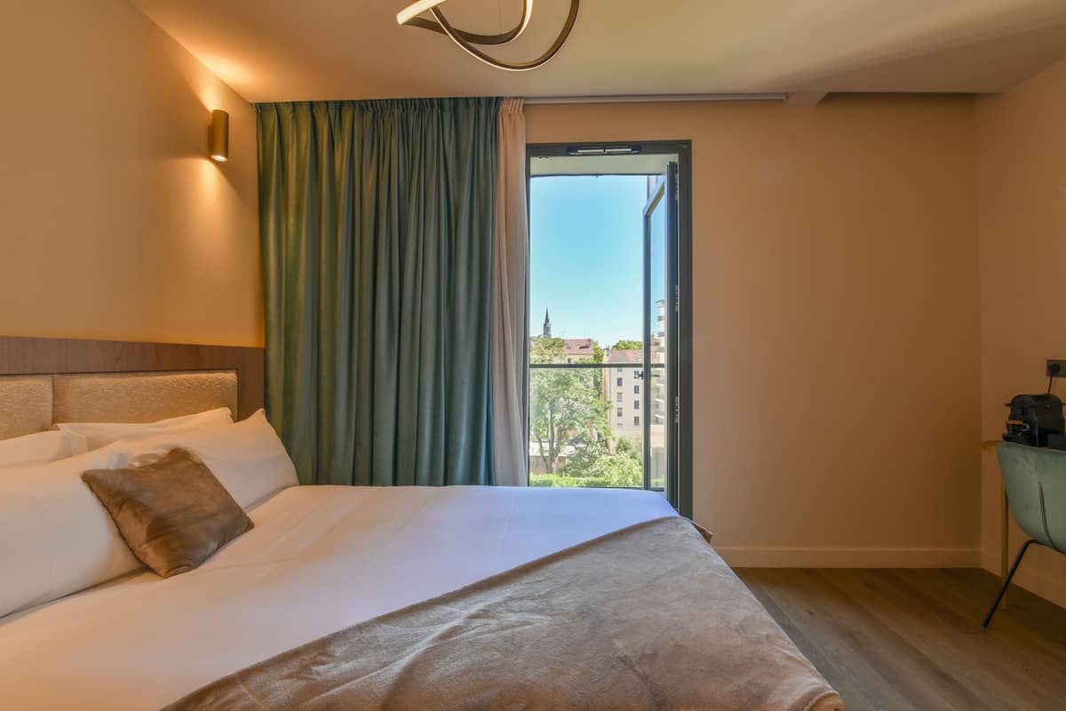 A hotel room with a bed, a nightstand, a lamp, a window with curtains, and a view of buildings outside.
